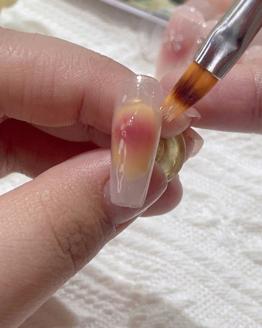 Un pincel pequeño difuminando suavemente el gel rosa sobre la superficie de la uña.