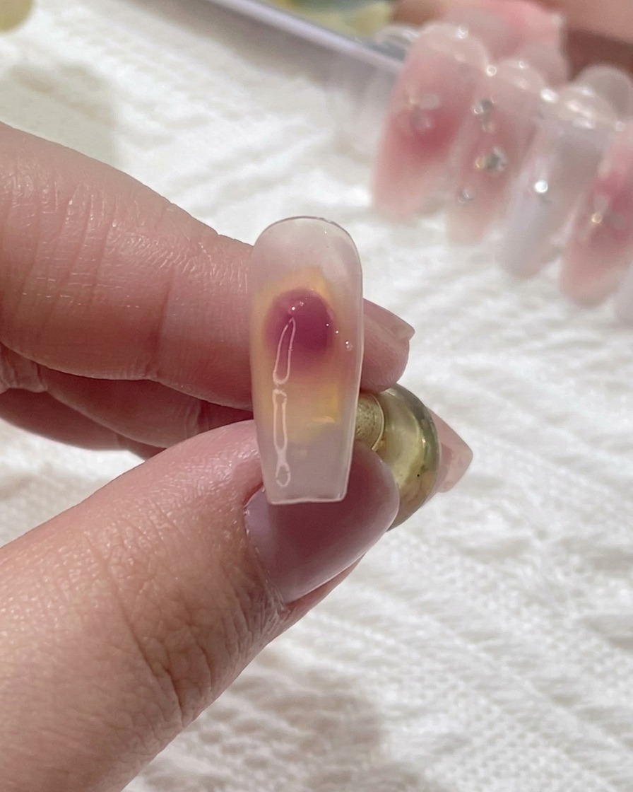 Primer plano de un pincel de uñas difuminando un punto de gel rosa sobre una uña transparente para crear un degradado blush suave.