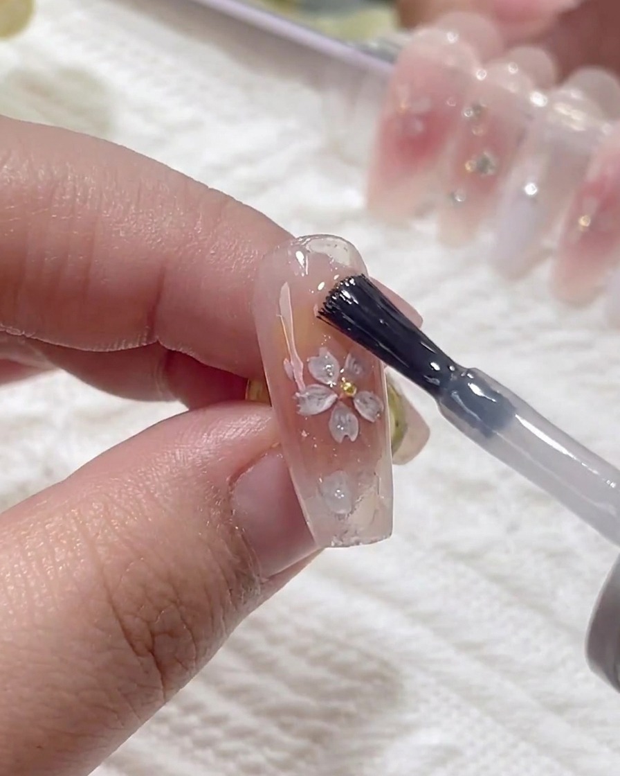 Un pincel aplicando una capa de gel de refuerzo transparente sobre la superficie de la uña, cubriendo las lentejuelas de flores de cerezo aplicadas.