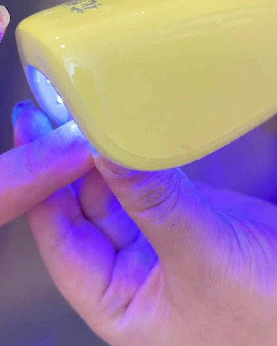 Una mano colocando una uña con gel blanco lechoso bajo una pequeña lámpara UV/LED para curar.