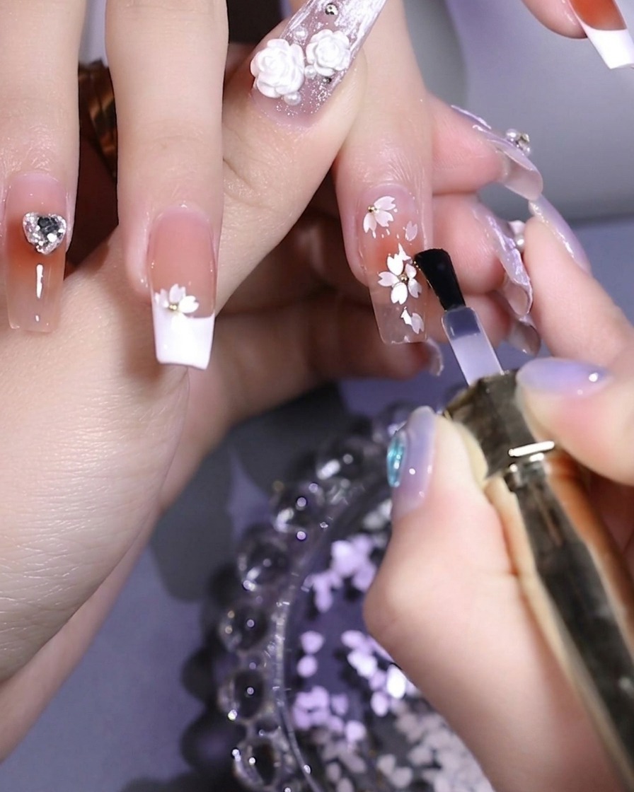 Un pincel aplicando un top coat transparente y brillante sobre una uña decorada con lentejuelas florales y una cuenta metálica central.