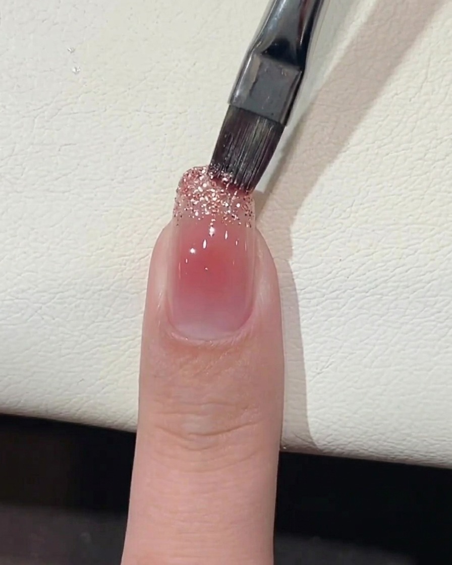 Un pincel aplicando gel con glitter grueso en el borde libre de la uña.