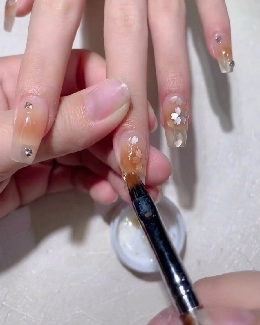 Primer plano de un pincel para arte de uñas alisando gel constructor sobre lentejuelas en forma de flor en una uña nude.