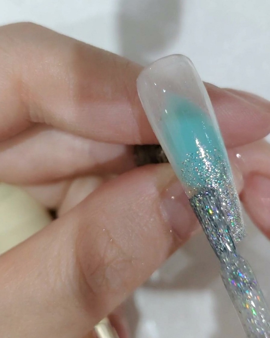 Primer plano de un pincel de uñas aplicando gel de purpurina plateada en la punta de una uña.