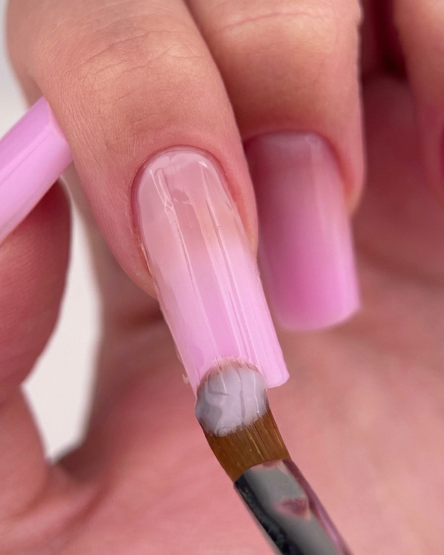 Un pincel aplicando un esmalte de gel rosa lechoso y claro cerca de la cutícula de la uña.