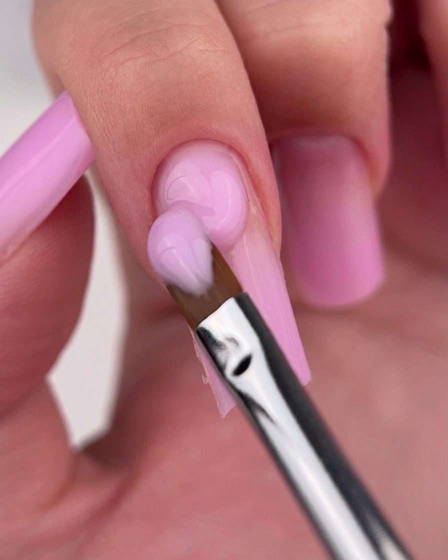 Un pincel de gel limpio difuminando suavemente la unión de dos tonos de esmalte rosa en una uña.