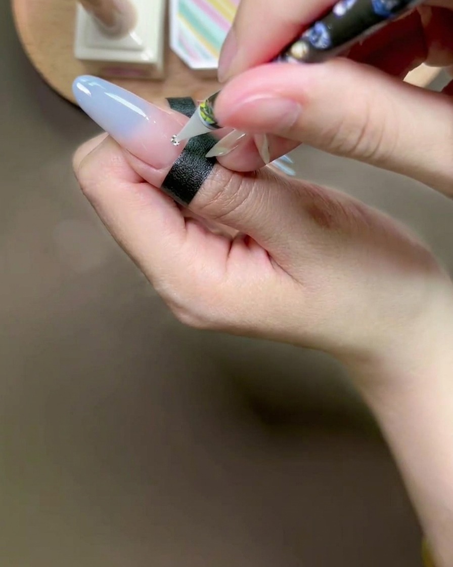 Un lápiz de cera colocando un pequeño cristal plateado sobre una gota de pegamento en una uña.