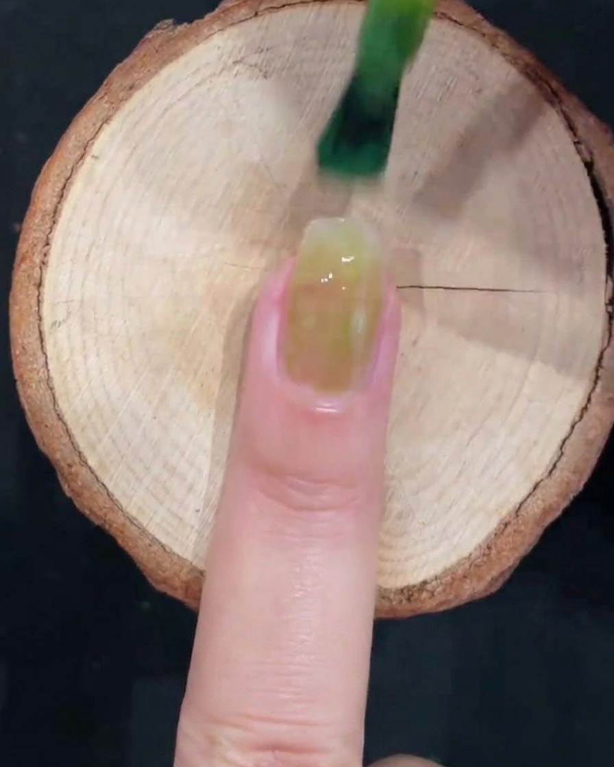 Un pincel de gel aplicando esmalte verde glacial translúcido en el centro de una uña natural limpia sobre una superficie de madera.