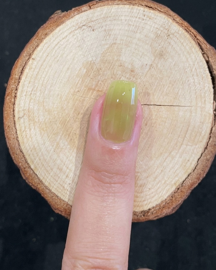 Primer plano de un pincel aplicando esmalte de gel verde translúcido sobre una uña natural.