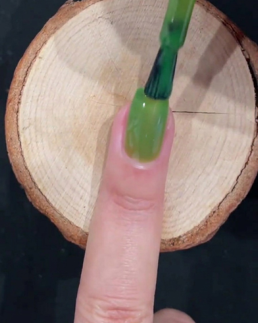 Un pincel de gel completando una capa completa de esmalte verde translúcido con trazos suaves a lo largo de la uña.