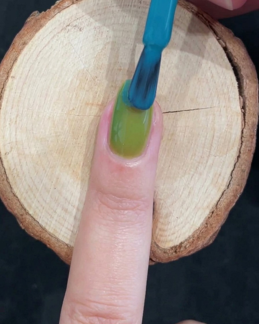 Un pincel de uñas aplicando un esmalte de gel azul vívido sobre la mitad superior de una uña pintada de verde brillante para crear un degradado de dos tonos.