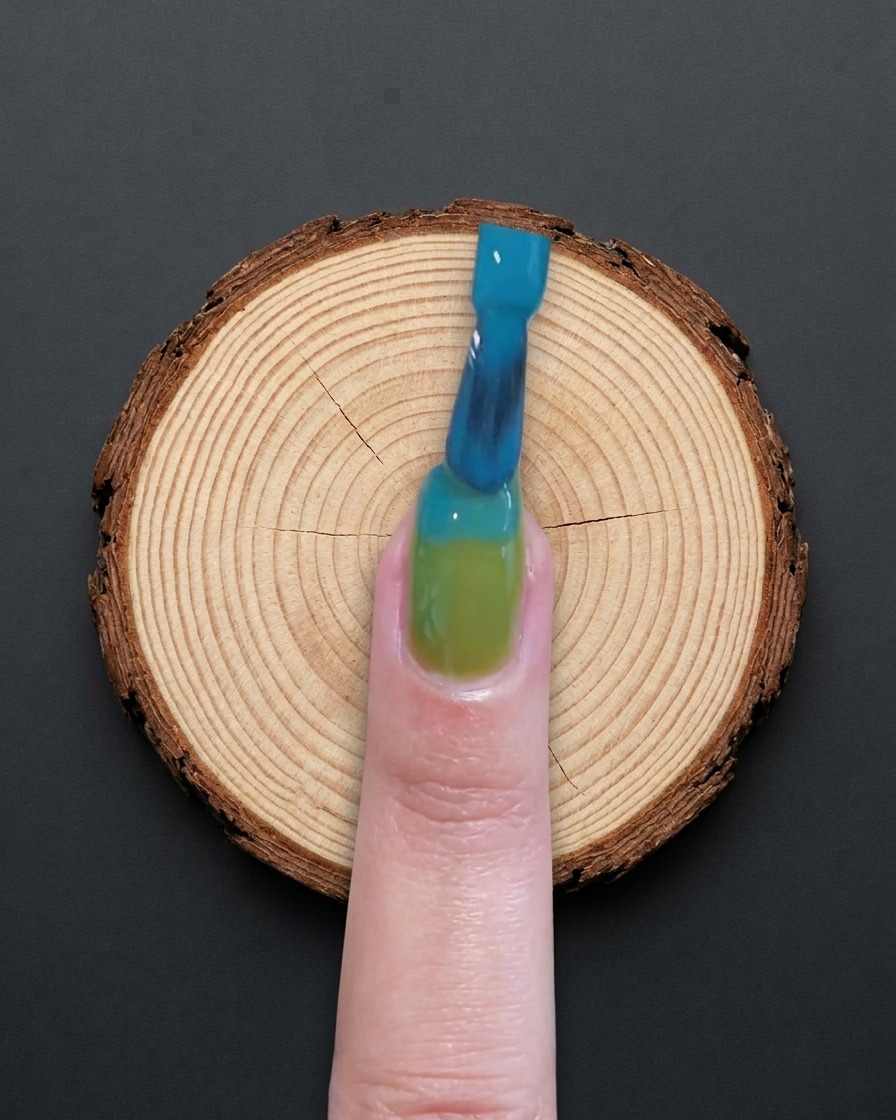 Un pincel aplicando esmalte de gel azul en la mitad superior de una uña con base verde.