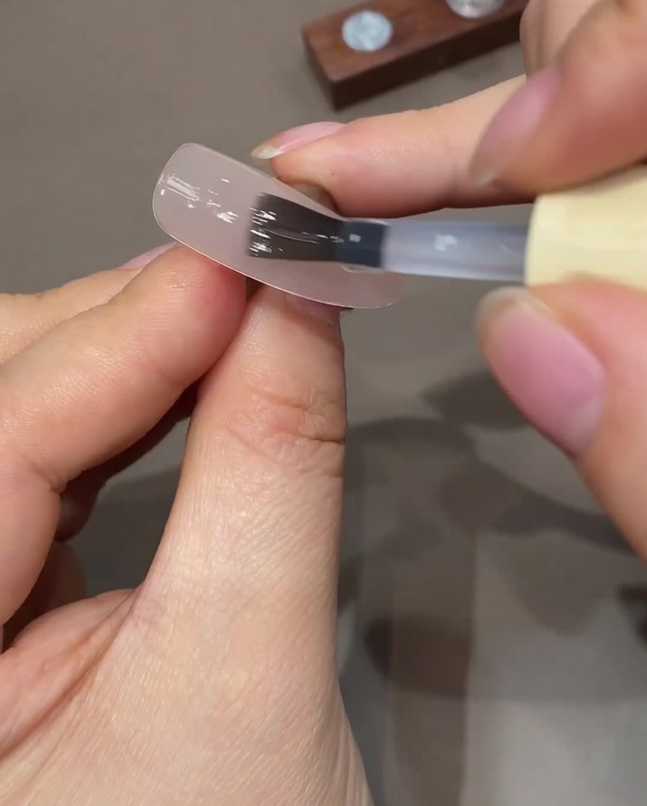 Un pincel de gel plano aplicando una capa fina y transparente de top coat sobre una uña nude preparada.