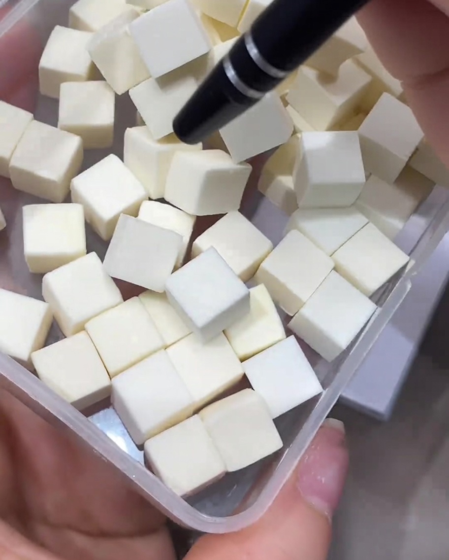 Una herramienta de precisión recogiendo un pequeño cubo de esponja de maquillaje blanca de un contenedor transparente lleno de cubos idénticos.