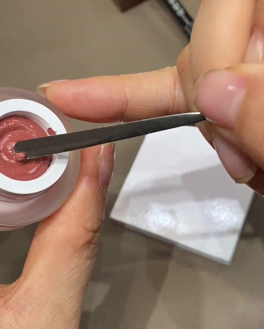 Una espátula metálica dispensando una pequeña gota de esmalte en gel de color sobre una paleta blanca.