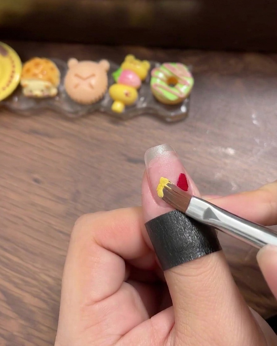 Primer plano de un pincel liner aplicando pequeños cuadrados definidos de esmalte en gel rojo, amarillo, azul y menta sobre un tip de uña transparente.
