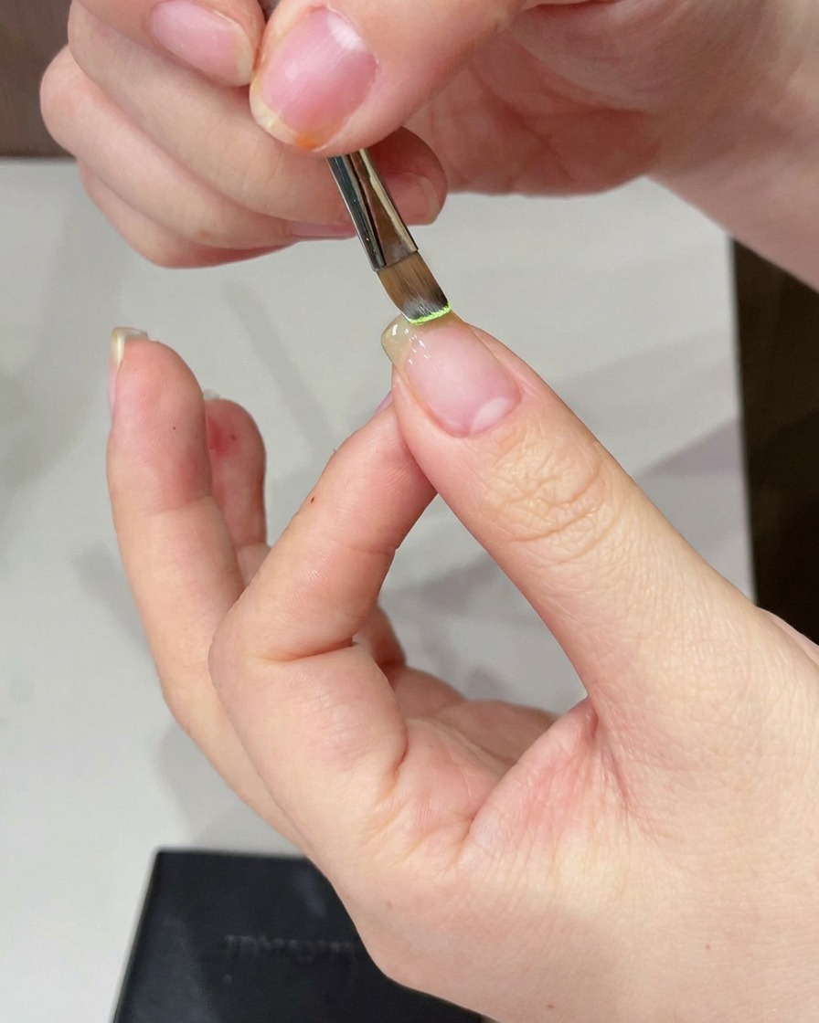 Primer plano de un pincel de detalle aplicando gel verde claro sobre la punta de una uña natural.
