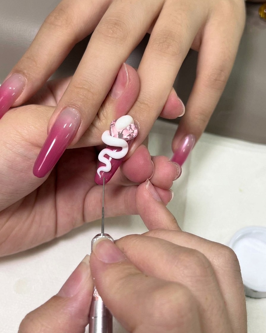 Primer plano de una mano usando una herramienta de metal de precisión para moldear una serpiente en gel 3D blanco sobre una gema rosa en una uña ombré magenta.