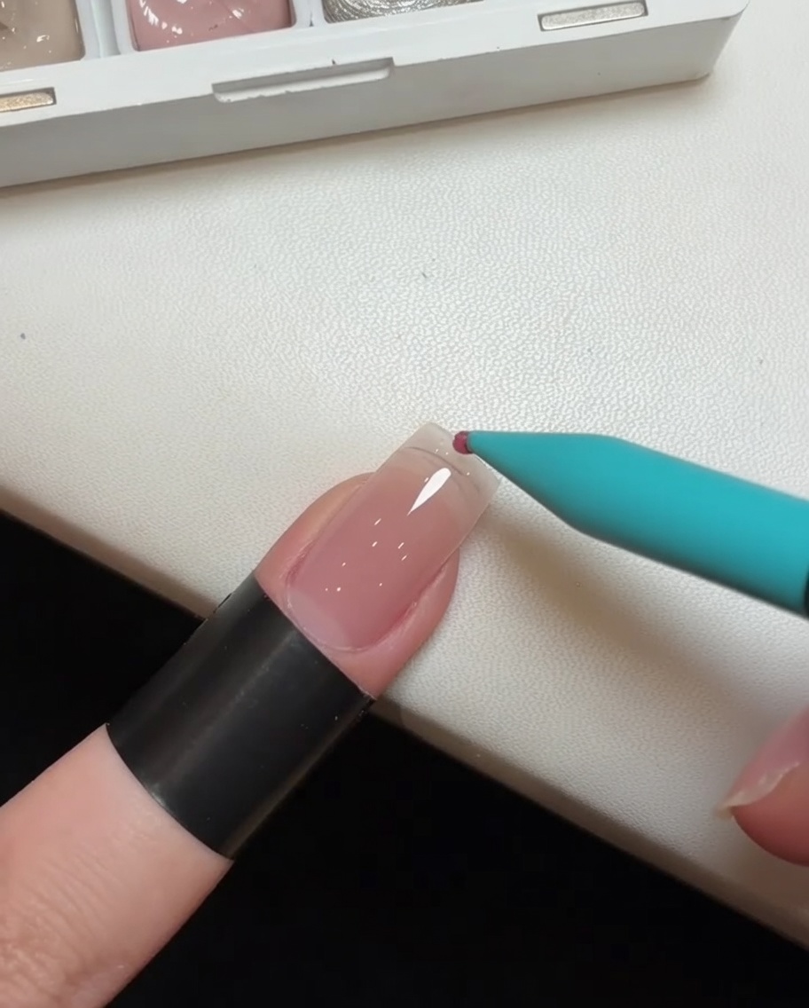 Primer plano de un pincel de detalle aplicando una gota de esmalte en gel rosa en la punta de una extensión de uña transparente.
