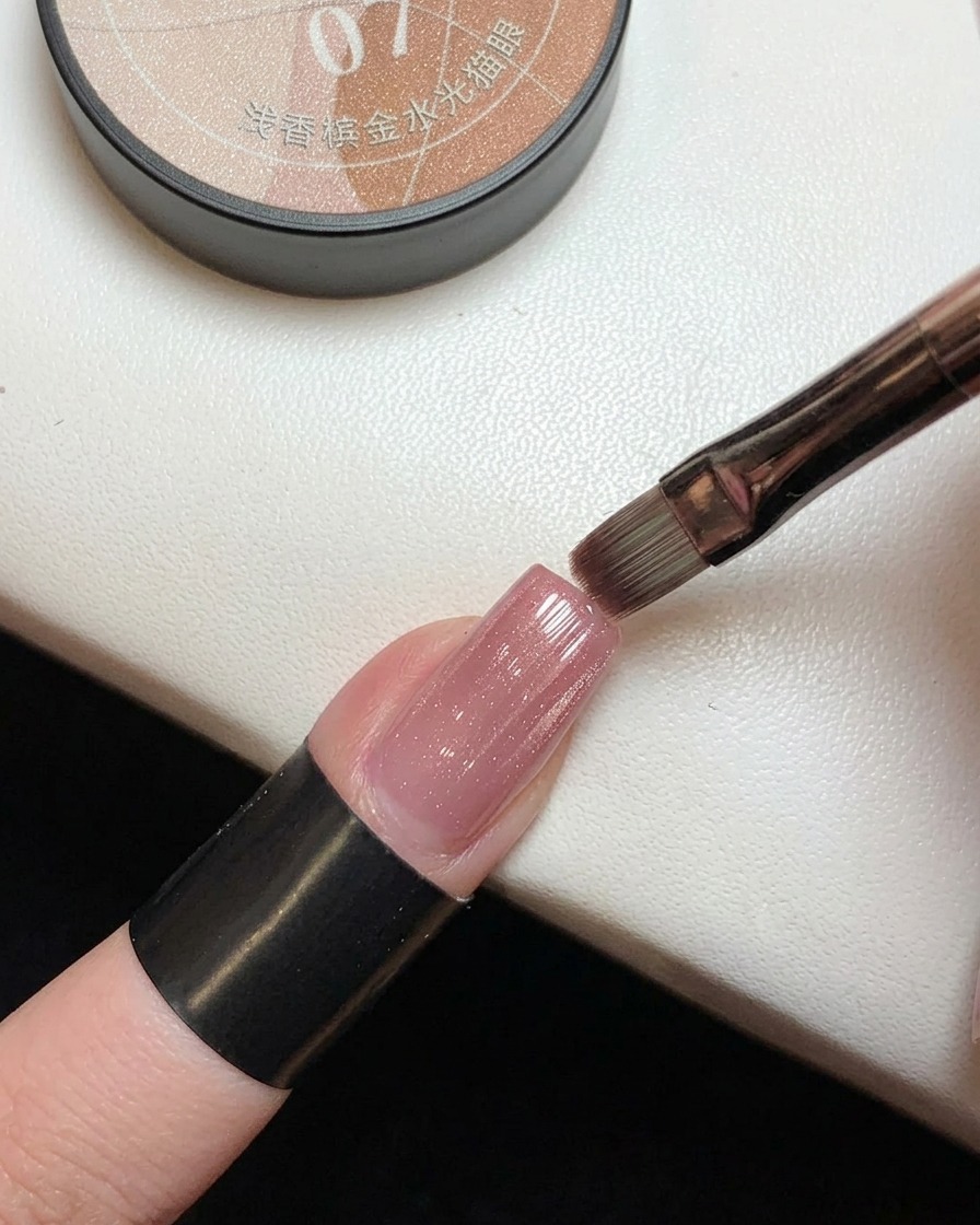 Un pincel aplicando gel magnético rosa translúcido con destellos dorados sobre una uña con degradado rosa previamente curada.