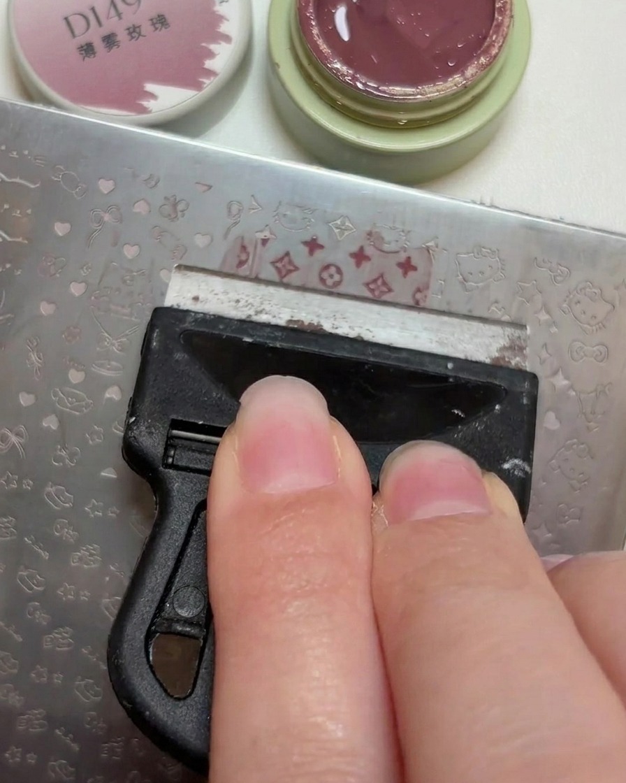 Un raspador de plástico nivelando una pequeña cantidad de esmalte en gel rosa empolvado a través de los patrones grabados en una placa de estampado de metal.