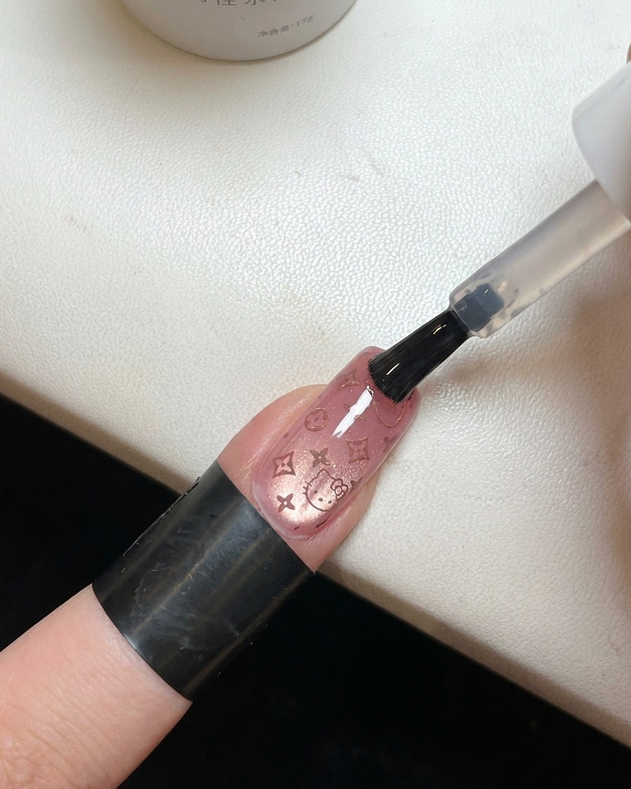 Un pincel aplicando un top coat transparente y brillante sobre una uña que tiene un patrón rosa de Hello Kitty.