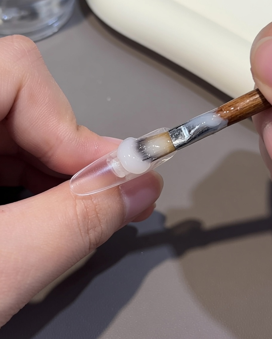 Primer plano de un pincel aplicando gel color nude en la mitad de la cutícula de un tip de uña transparente.