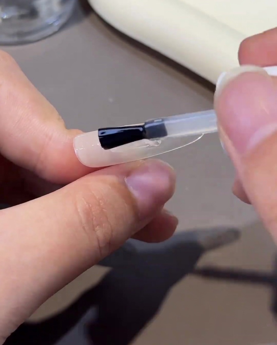 Un pincel aplicando un gel fortalecedor transparente y viscoso sobre la uña, creando una superficie lisa.