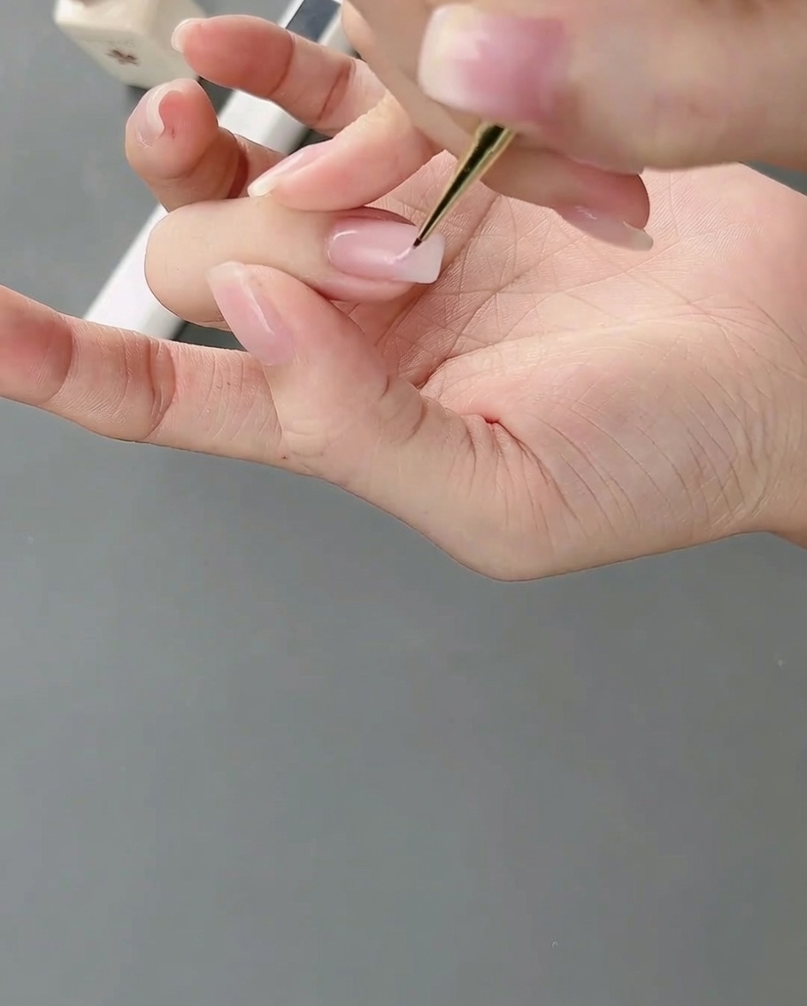 Un pincel de detalle fino aplicando gel rosa claro sobre una uña para formar bloques de color irregulares.