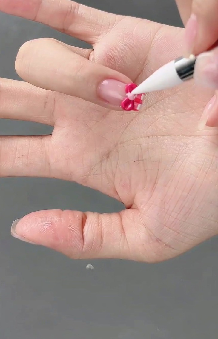 Una pequeña perla de gel rosa claro siendo colocada en el centro de un lazo 3D usando un pincel de detalle.