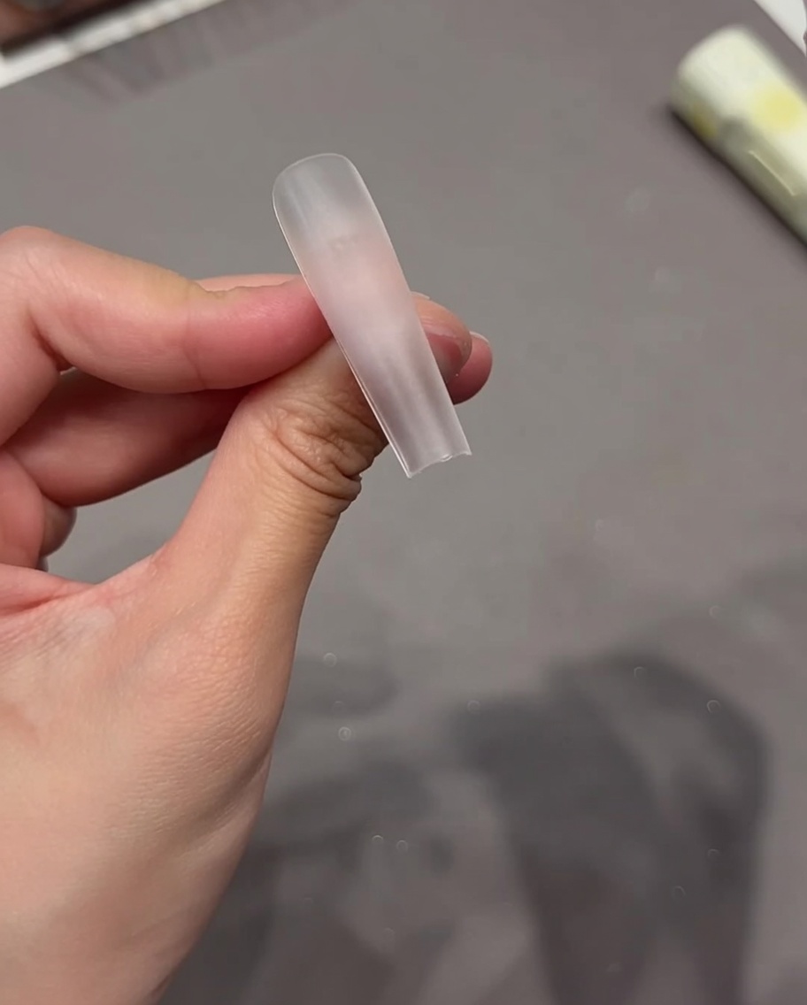 Primer plano de una mano aplicando una pegatina adhesiva transparente sobre la superficie de una punta de uña clara.