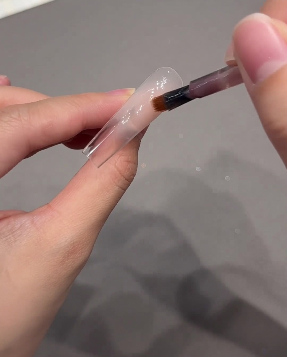 Un pincel de arte de uñas aplicando un punto de gel rosa nude en el área de la cutícula de la punta de la uña.