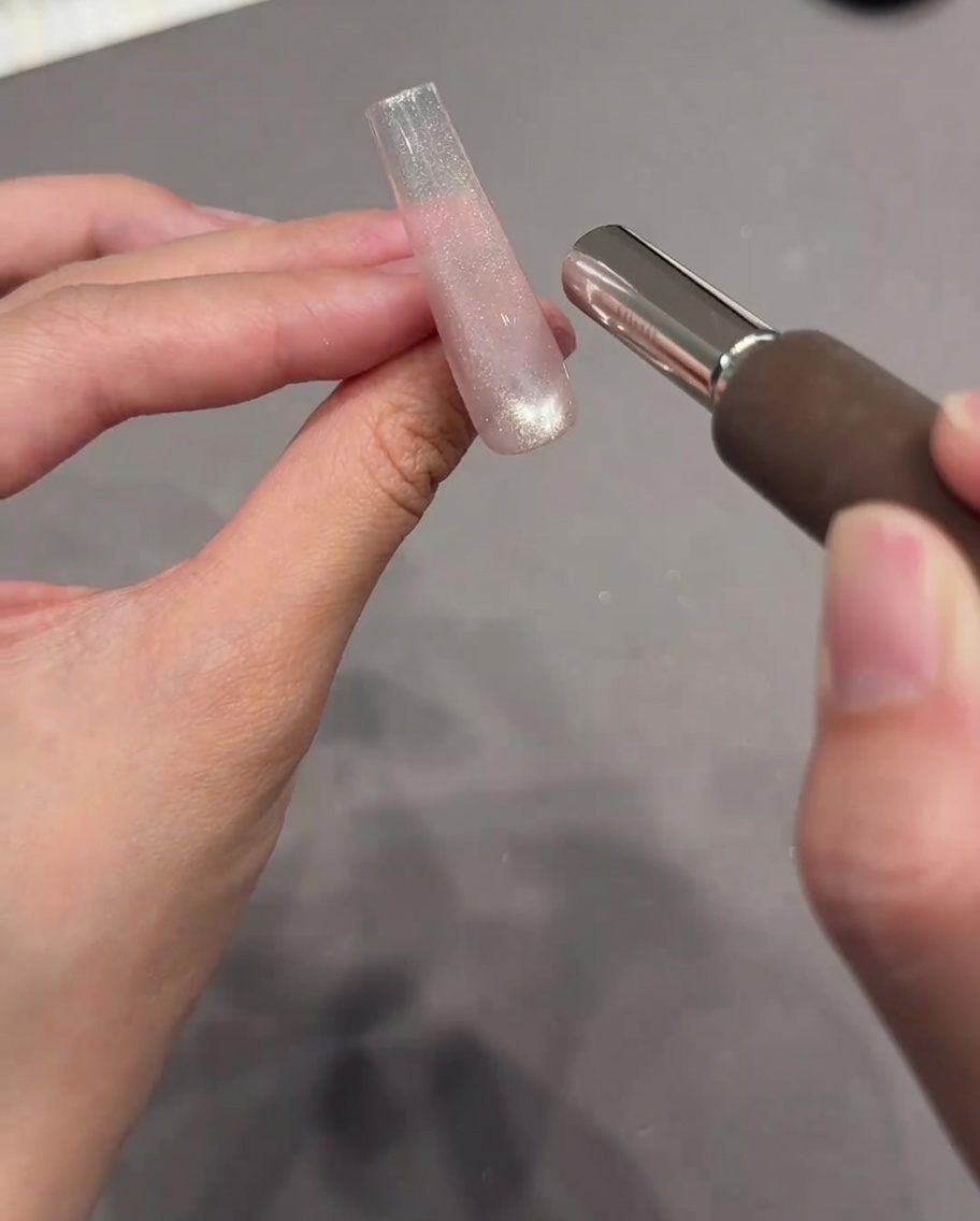 Una mano sosteniendo una herramienta magnética cilíndrica cerca de una extensión de uña transparente recubierta con gel ojo de gato brillante.