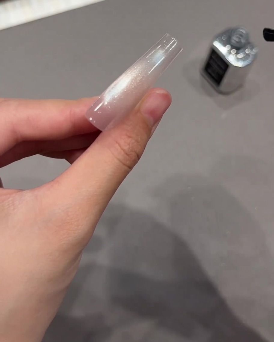 Un pincel de uñas negro aplicando un top coat transparente sobre una extensión de uña de gel ojo de gato brillante ya curada.