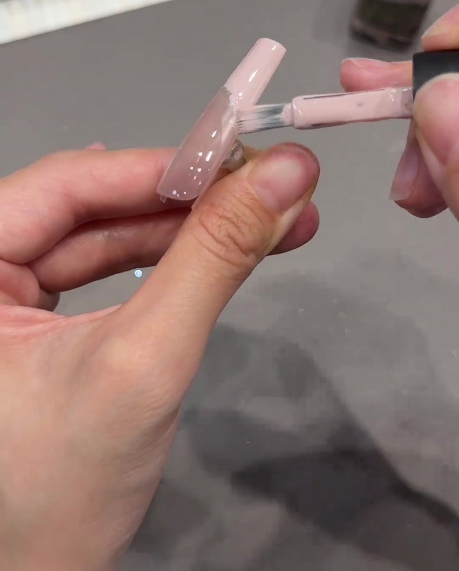 Un pincel de uñas aplicando gel rosa nude opaco en la punta de una uña, usando una pegatina transparente curva como límite.