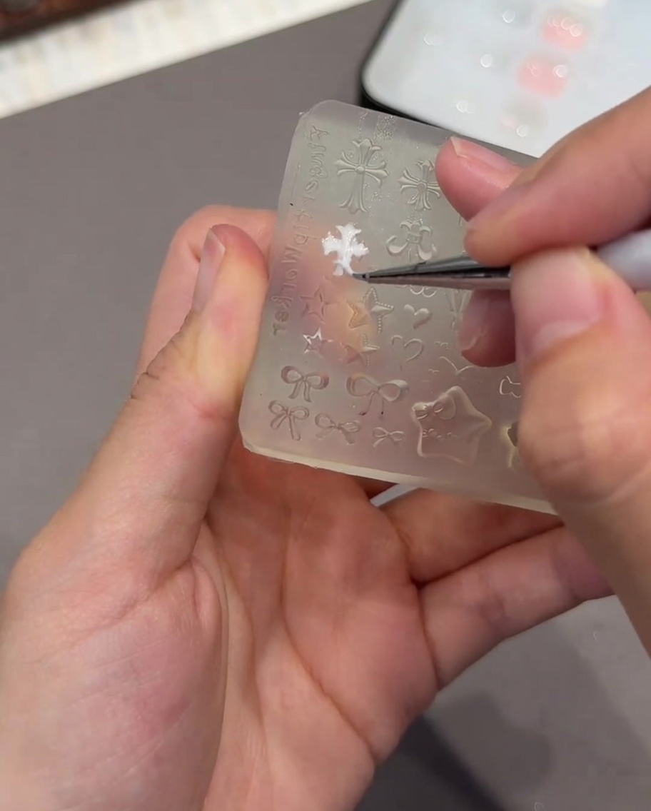 Un par de pinzas sosteniendo un pequeño molde 3D de corazón blanco, preparándose para colocarlo sobre una uña.