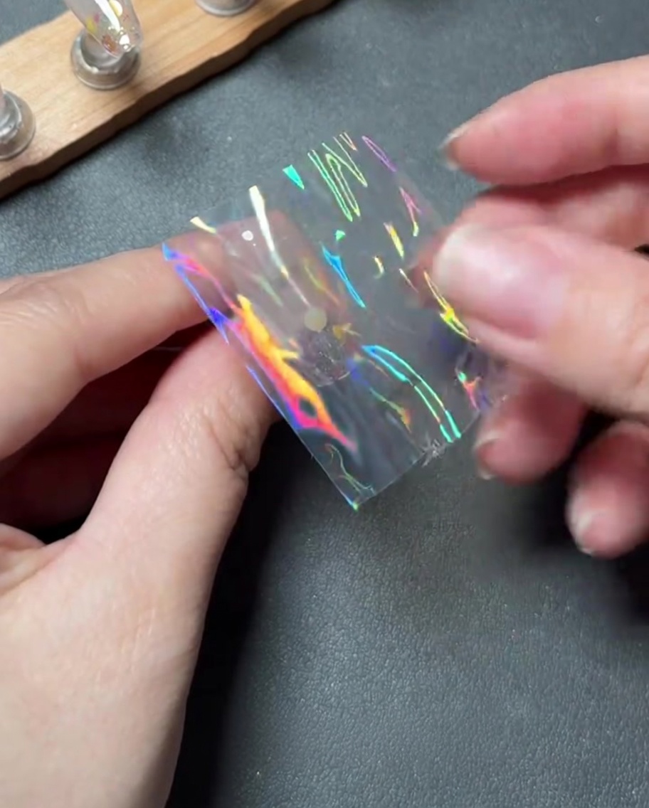 Dedos colocando cuidadosamente un pequeño trozo de foil de transferencia holográfico en el centro de una uña.