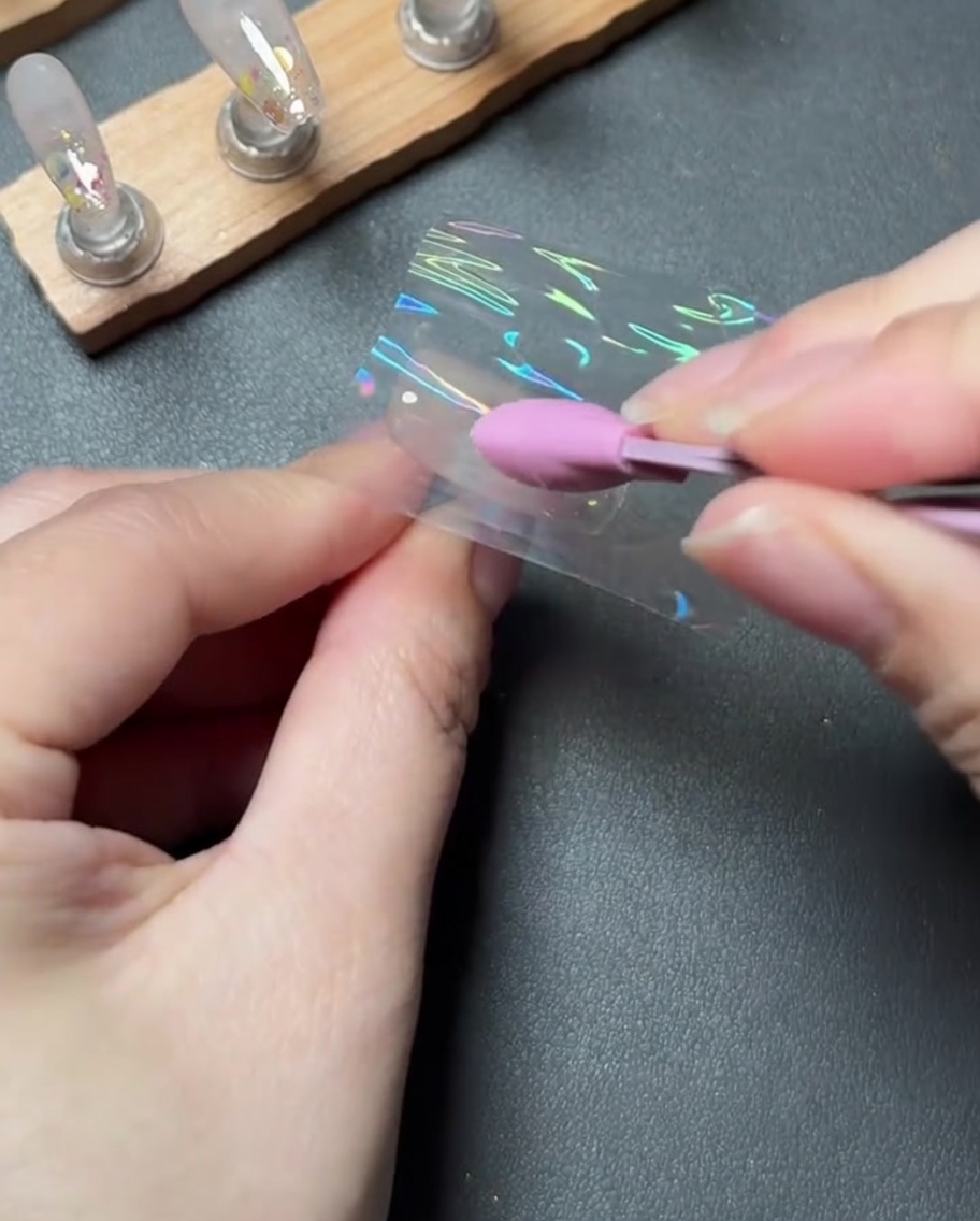 Una herramienta de silicona rosa presionando y alisando firmemente un trozo de foil de transferencia holográfico sobre una uña.