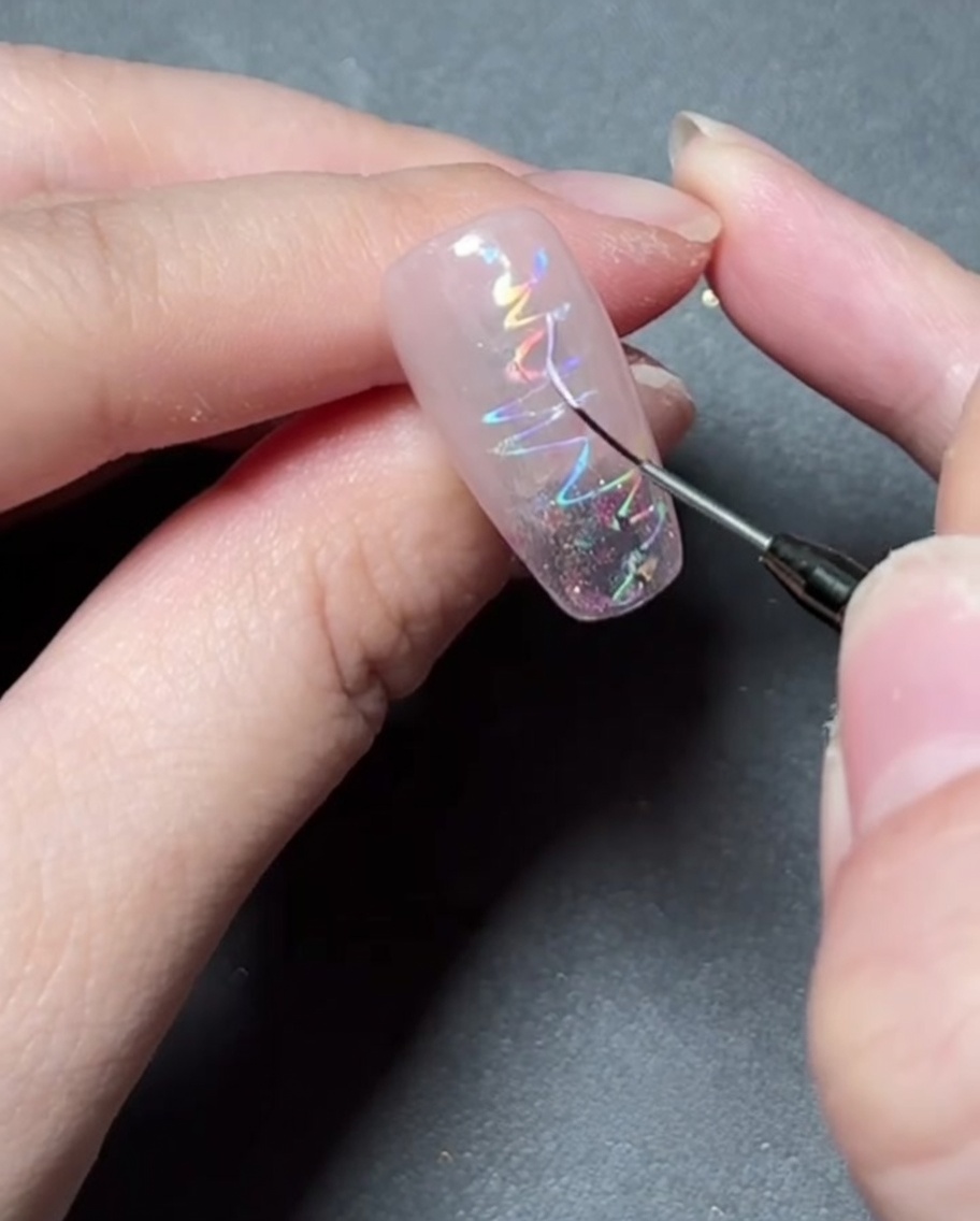 Un pincel delineador fino pintando una delicada forma de estrella con gel colorido sobre una uña con foil holográfico.