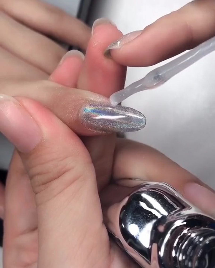 Un pincel aplicando gel de refuerzo transparente sobre una uña cromada, cubriendo el borde libre.