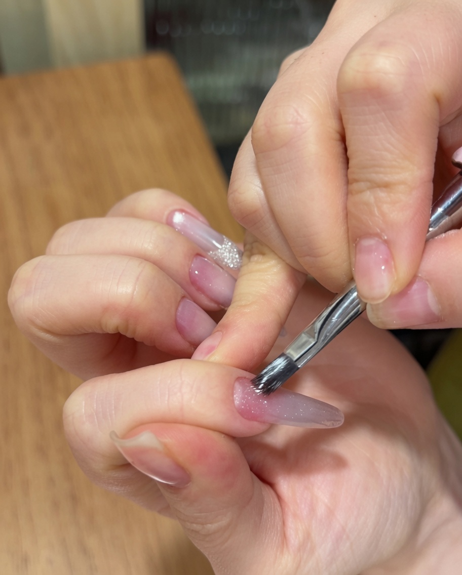 Una mano usando un pincel para uñas para aplicar un esmalte en gel cat eye brillante sobre una uña almendrada larga con una base rosa degradada.
