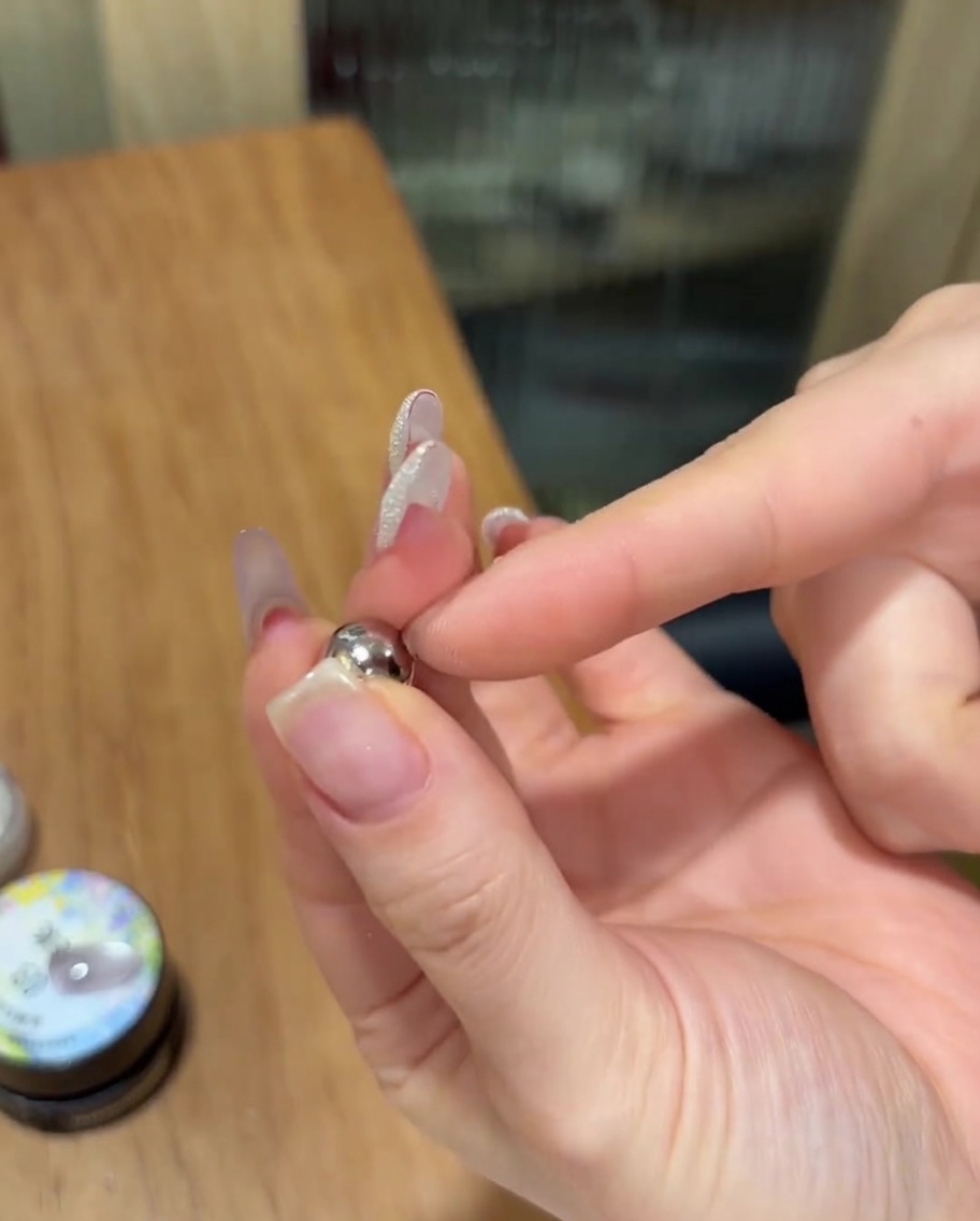 Una mano sosteniendo un pequeño imán esférico plateado cerca de una uña pintada con gel cat eye brillante sin curar.