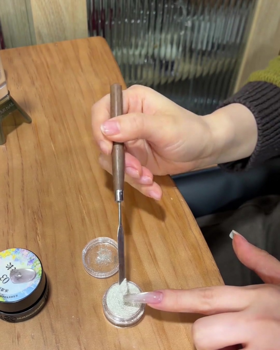 Un primer plano de una uña con gel transparente sin curar siendo cubierta con fino polvo de azúcar blanco usando una espátula de metal.