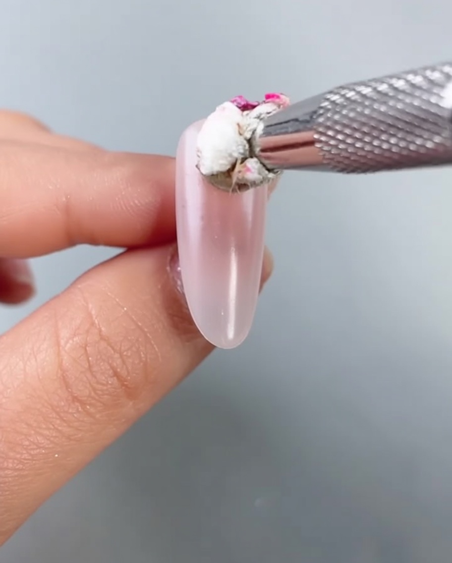 Un primer plano de una pequeña esponja aplicando esmalte de gel rosa brillante en el centro de una uña almendrada para crear un efecto de rubor difuminado.
