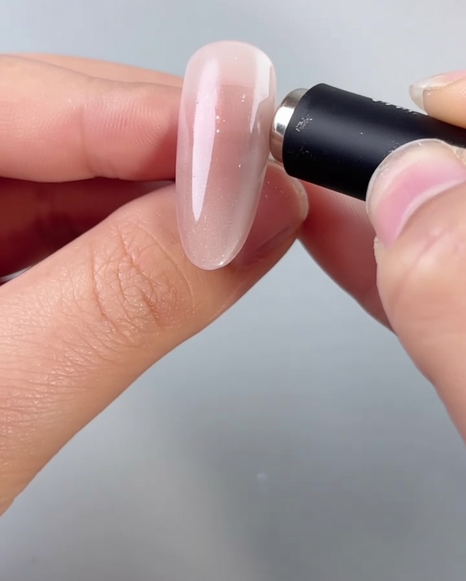 Una mano sosteniendo un imán cilíndrico cerca de una uña almendrada, activando las partículas brillantes en el gel ojo de gato húmedo.