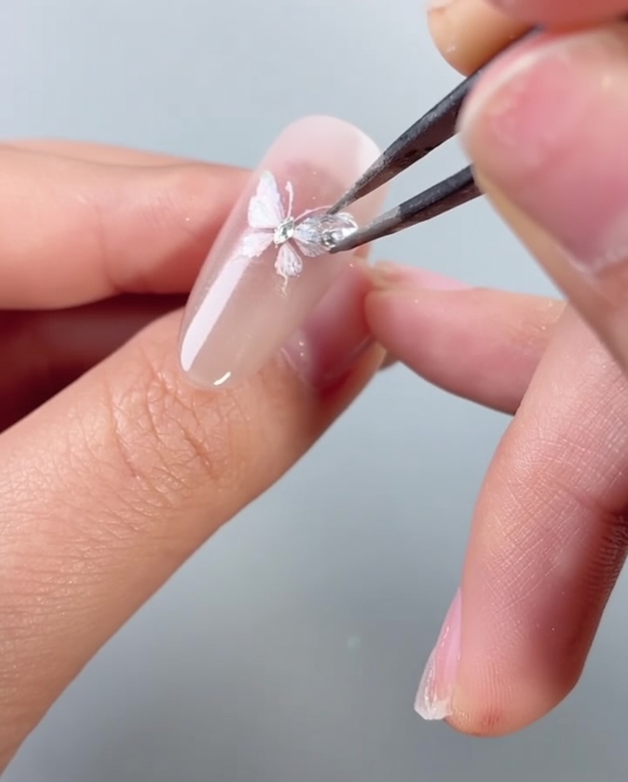 Unas pinzas colocando una pequeña piedra de diamante brillante sobre un diseño de arte de uñas de mariposa.