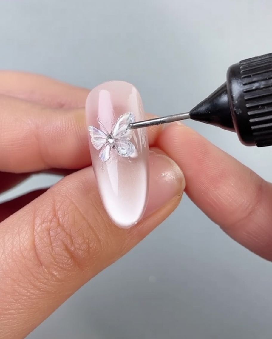 Un pincel aplicando una fina capa de gel transparente alrededor de la base de una decoración de mariposa en una uña.