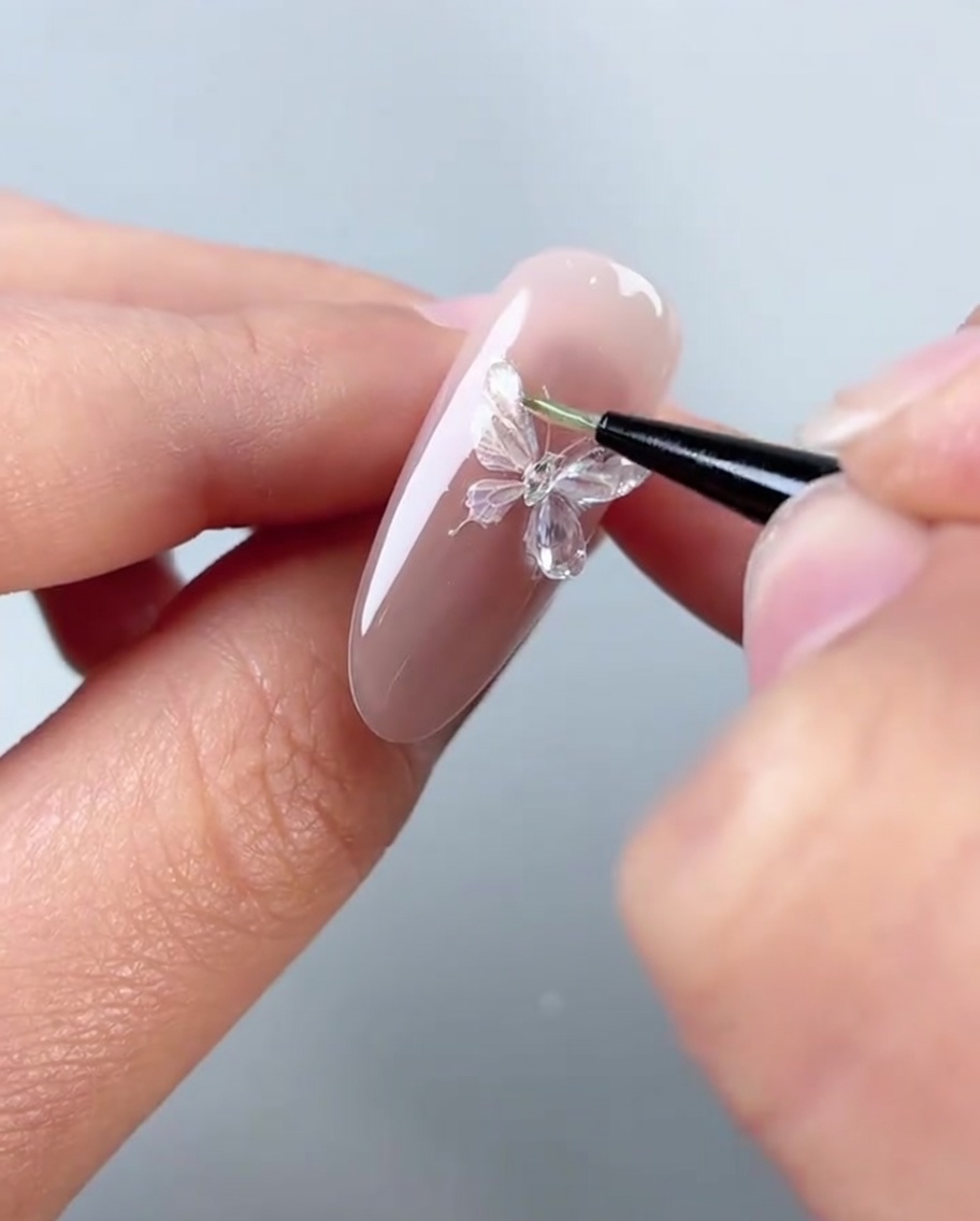 Una vista de cerca de un pincel aplicando pegamento de modelado transparente y espeso sobre las alas izquierdas de una pegatina de mariposa en una uña rosa.