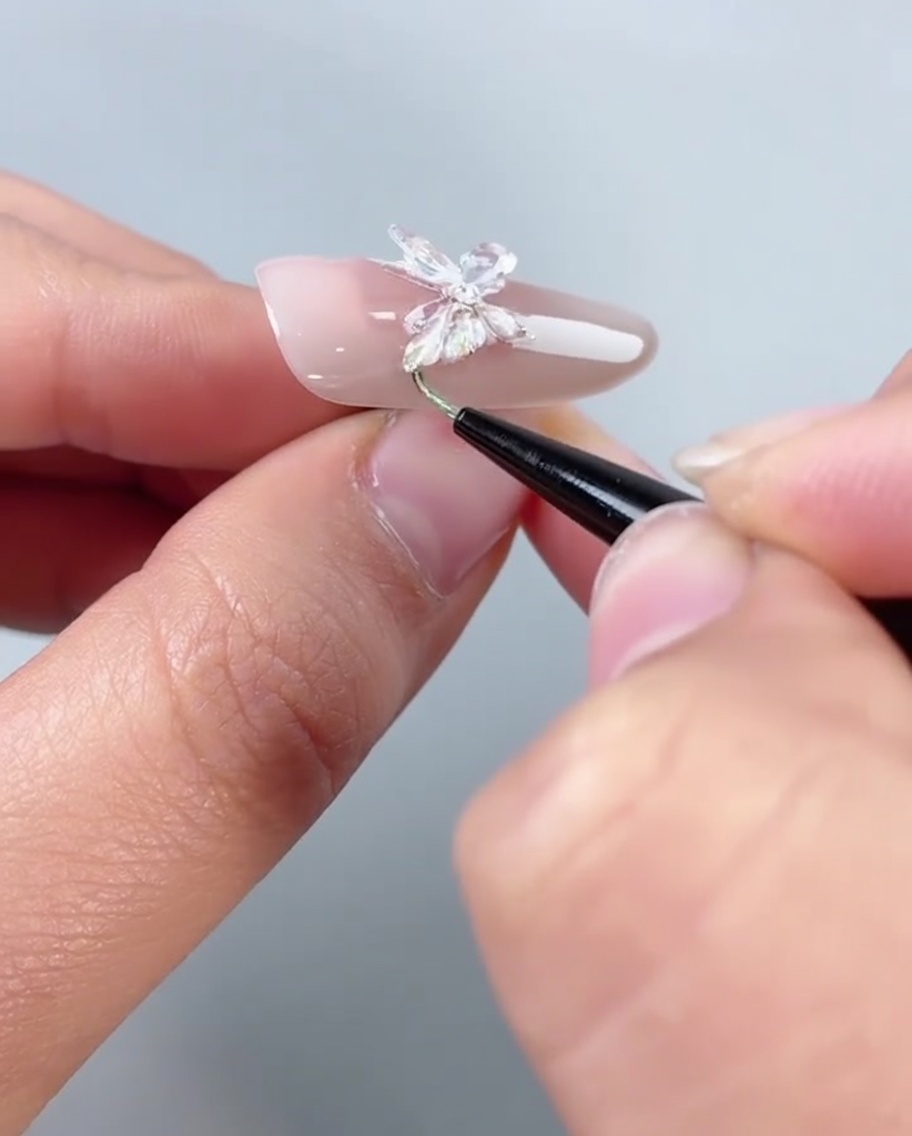 Un pincel aplicando pegamento de relleno transparente a lo largo del borde inferior de una uña decorada con una mariposa.