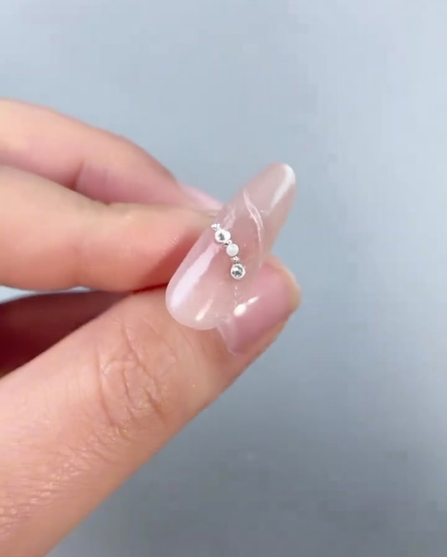 Un primer plano de un encanto de pentagrama plateado siendo colocado sobre una uña decorada con pequeñas perlas y cuentas.
